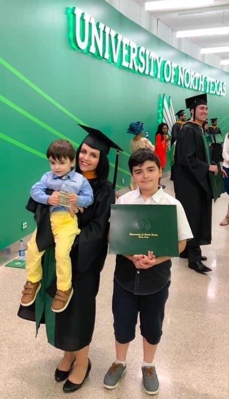 Nella in her cap and gown from UNT standing next to her oldest son. The oldest son is holding her degree, and she is holding her younger son.